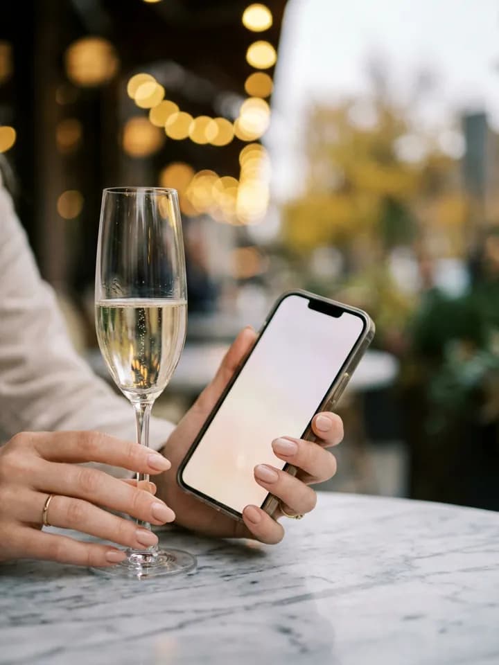 Mão segurando o celular ao lado de uma taça de champanhe